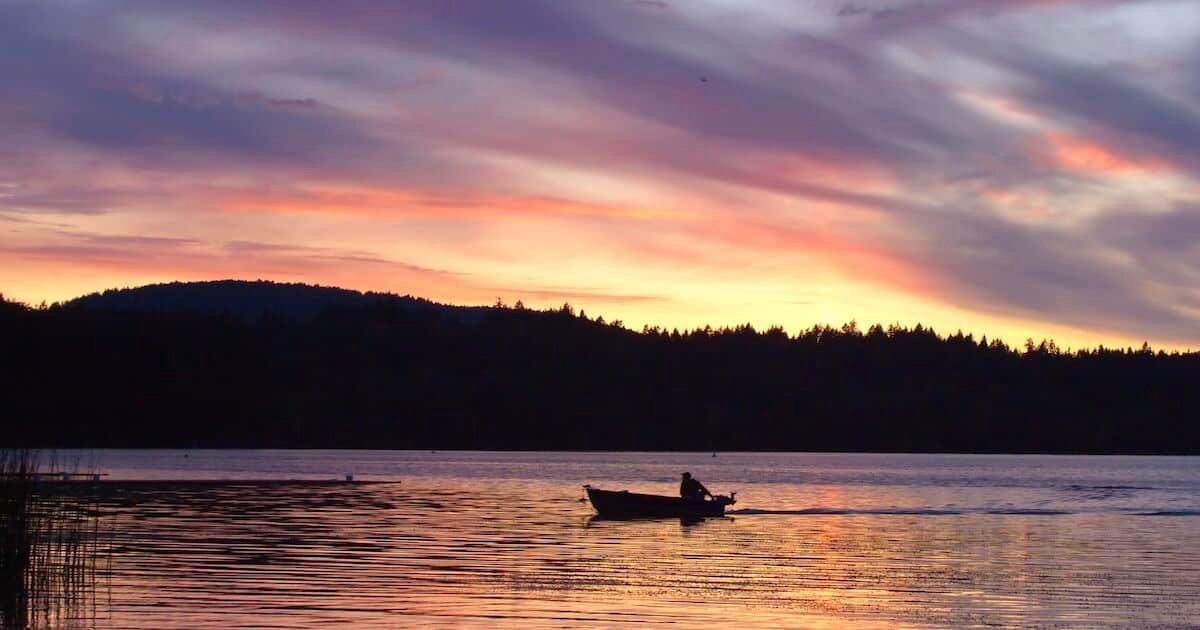 Silhouette of a small boat on a lake during sunset, with colorful clouds reflecting on the water's surface.