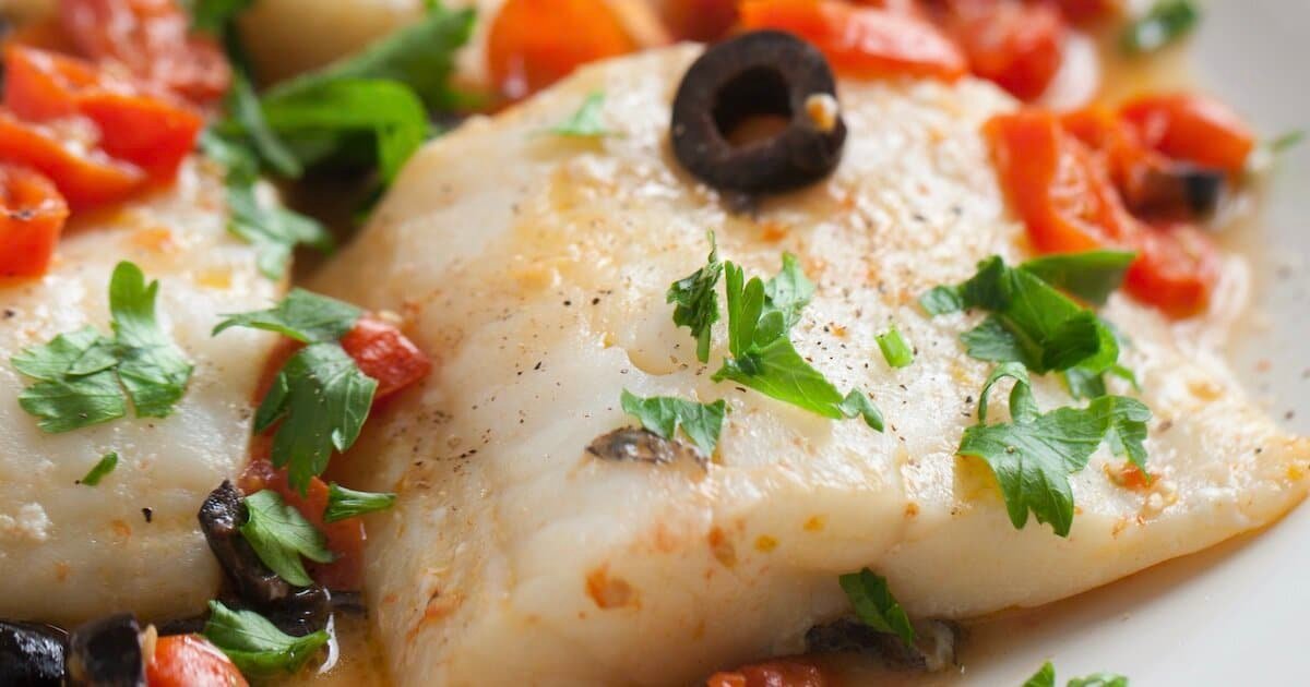 Cooked halibut fillet garnished with olives, tomatoes, and fresh herbs on a plate.