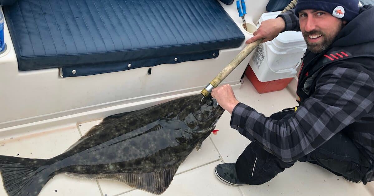 Person holding a large halibut on a boat deck, with fishing gear and equipment visible in the background.