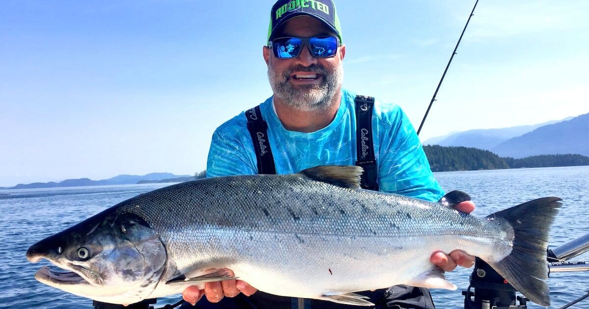 Man holding a large Coho salmon while fishing on a boat in a scenic water setting.