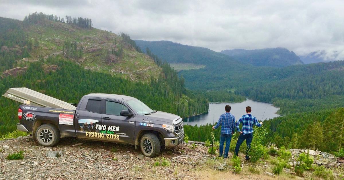 Two men standing beside a pickup truck with a fishing rods logo, overlooking a forested landscape and a lake in the distance.
