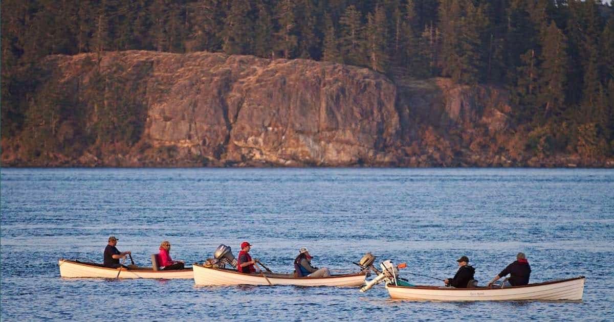 Four small boats with individuals fishing in a calm body of water, surrounded by forested cliffs in the background.