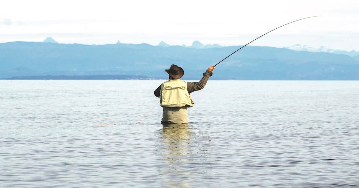 Person casting a fishing line while standing in shallow water, with mountains visible in the background.
