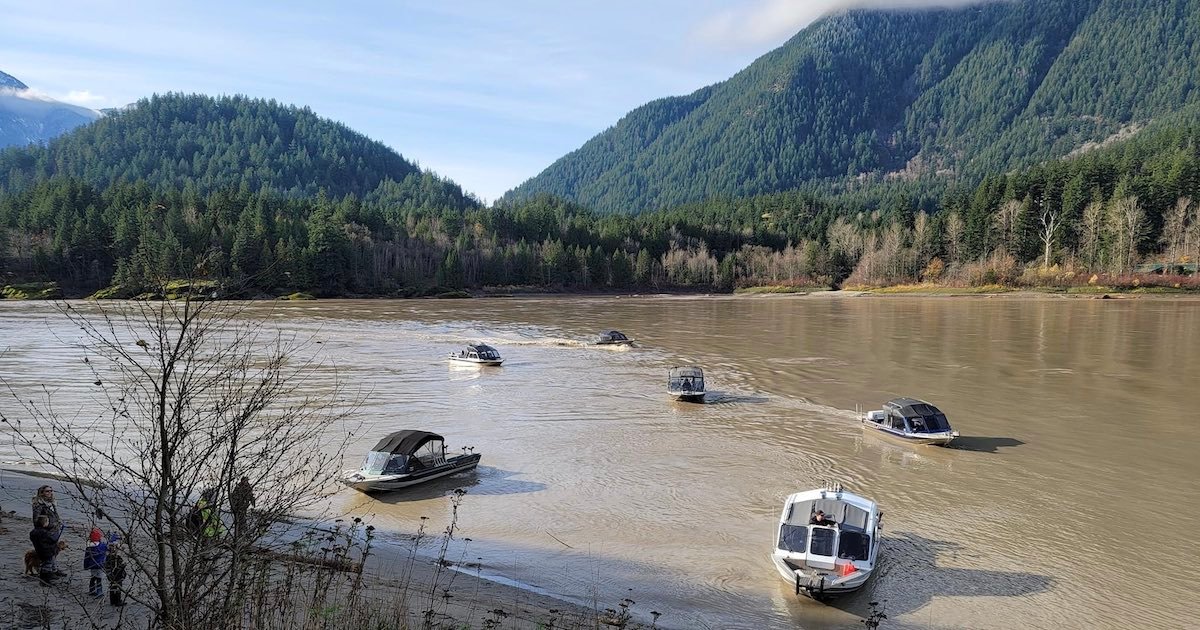 Multiple boats navigating through a river surrounded by forested mountains on a clear day.