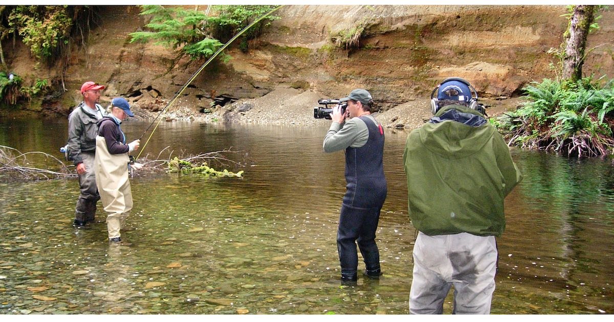 Two individuals fishing in a river while a third person records the activity with a camera.