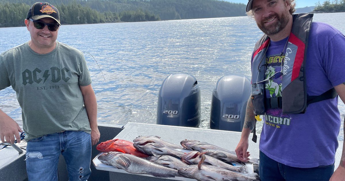 Two men on a boat displaying a catch of various fish, including lingcod, on a table in front of them.