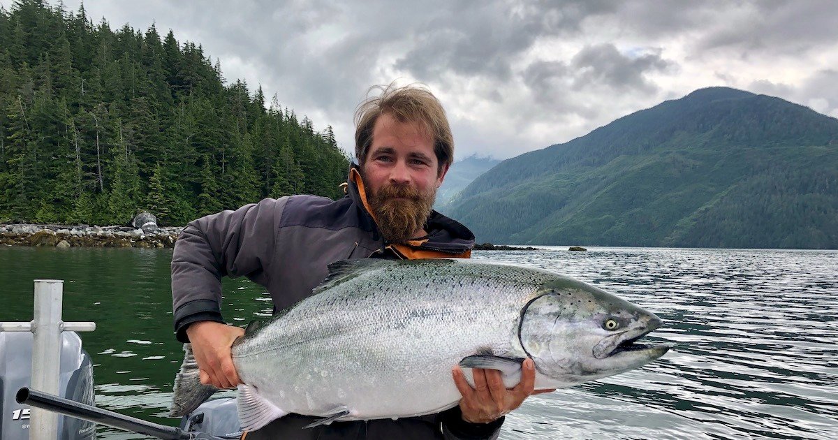 Man holding a large salmon fish while standing on a boat in a natural water setting with mountains and trees in the background.