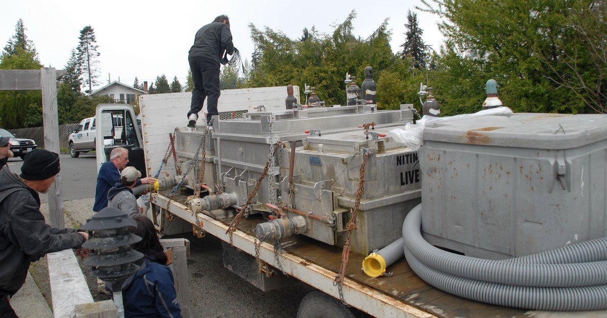 Transport of smolt tanks from Nitinat hatchery on a flatbed truck with workers securing the equipment.
