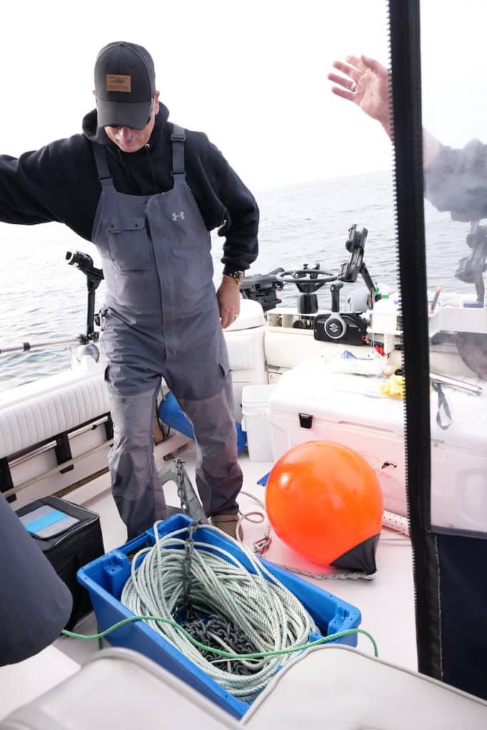 An angler preparing a halibut anchoring setup on a boat deck, featuring a coiled floating mainline and heavy anchor chain in a blue storage bin alongside a large orange Scotchman buoy used for safe retrieval in BC waters.
