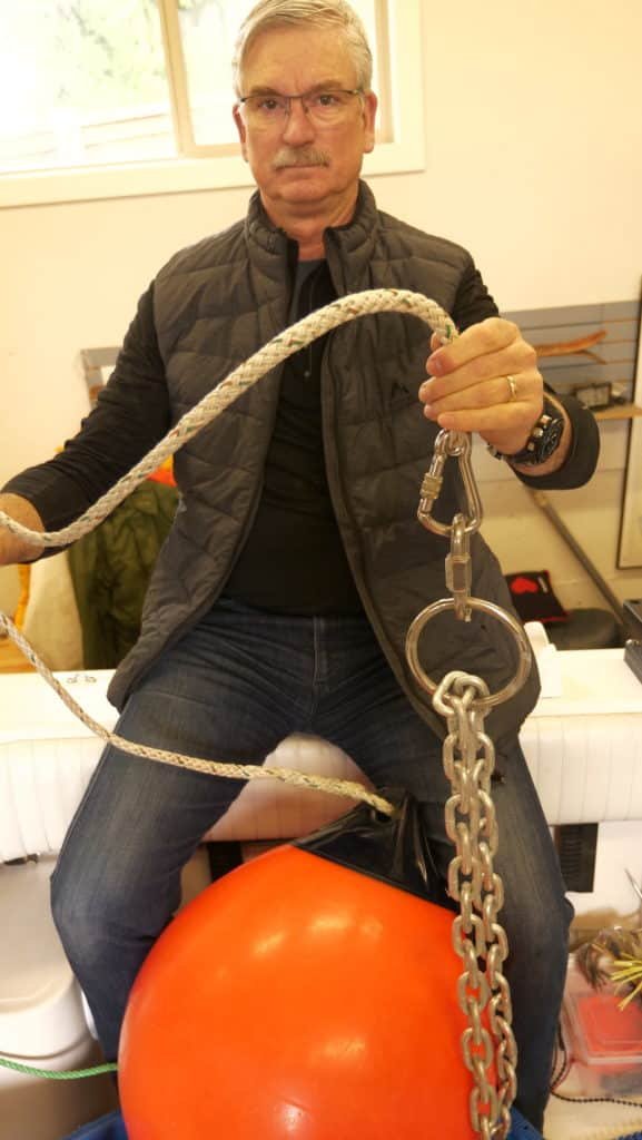 A demonstration of a halibut anchoring retrieval system featuring a large orange Scotchman buoy, a heavy-duty anchor chain, and a sliding ring with a locking carabiner used for safe anchor lifting in BC waters.