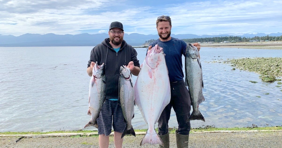 Two men holding large halibut and salmon caught during a fishing trip, standing on a rocky shoreline with water in the background.