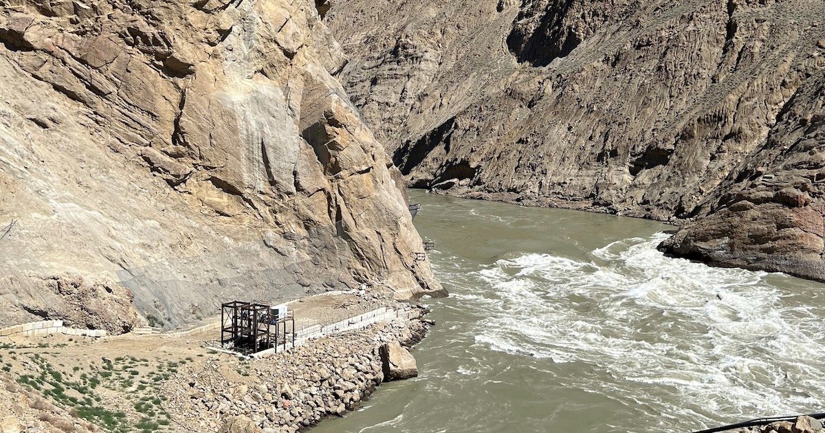 Rocky riverbank with a flowing river and a small structure near the water's edge, surrounded by steep cliffs.