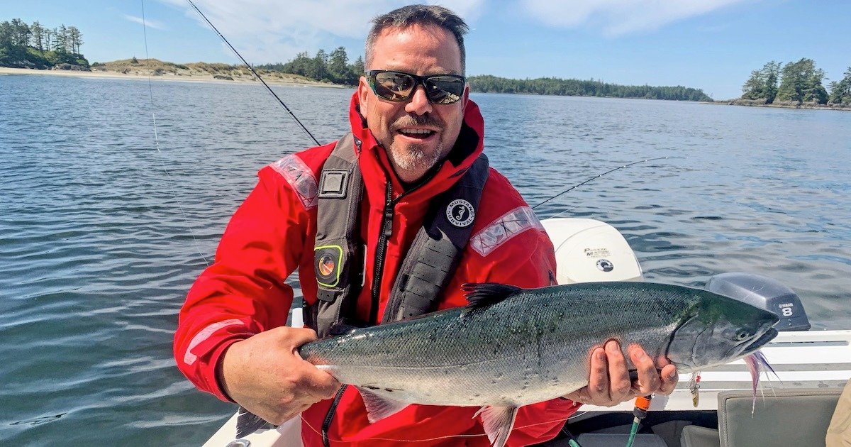Man in a red jacket holding a caught fish while standing on a boat in a body of water.