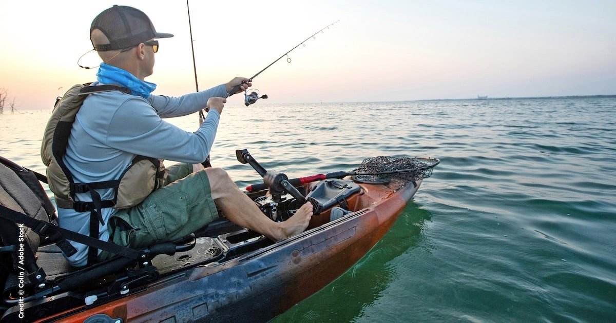 Person fishing from a kayak in calm waters during sunset, wearing a cap and fishing gear.