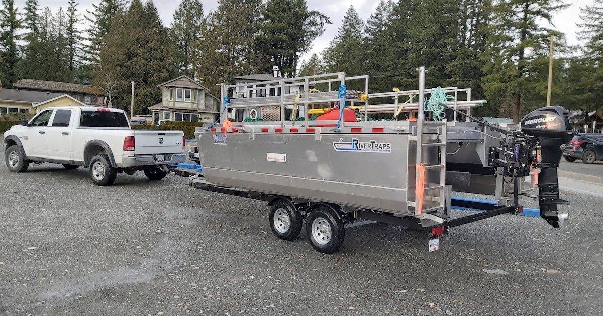 Silver river trap trailer attached to a white pickup truck, designed for fishing activities, parked on a gravel surface.