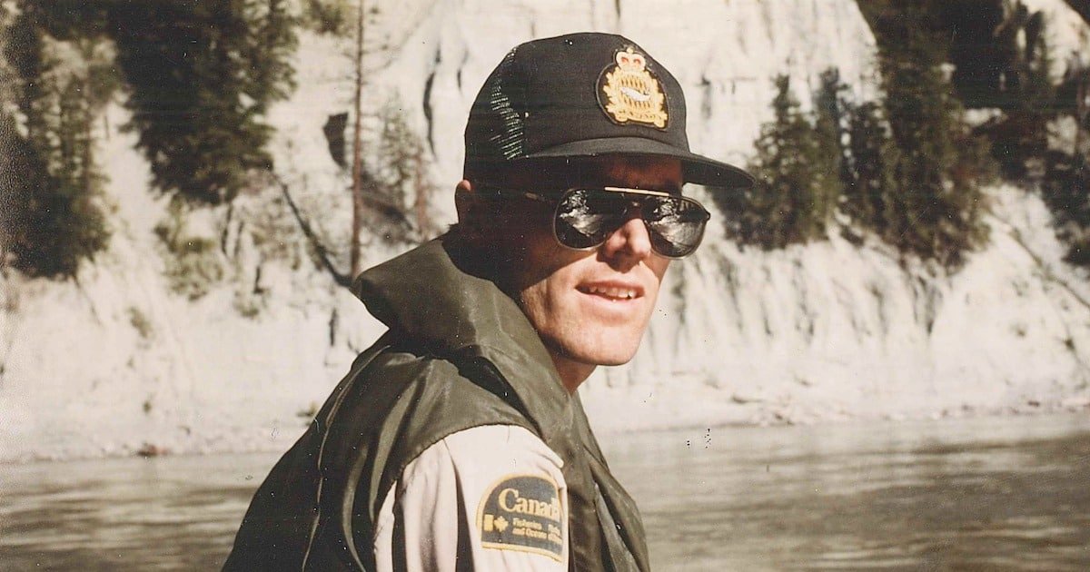 Man wearing sunglasses and a cap with a badge, dressed in a fishing vest near a riverbank with rocky cliffs in the background.