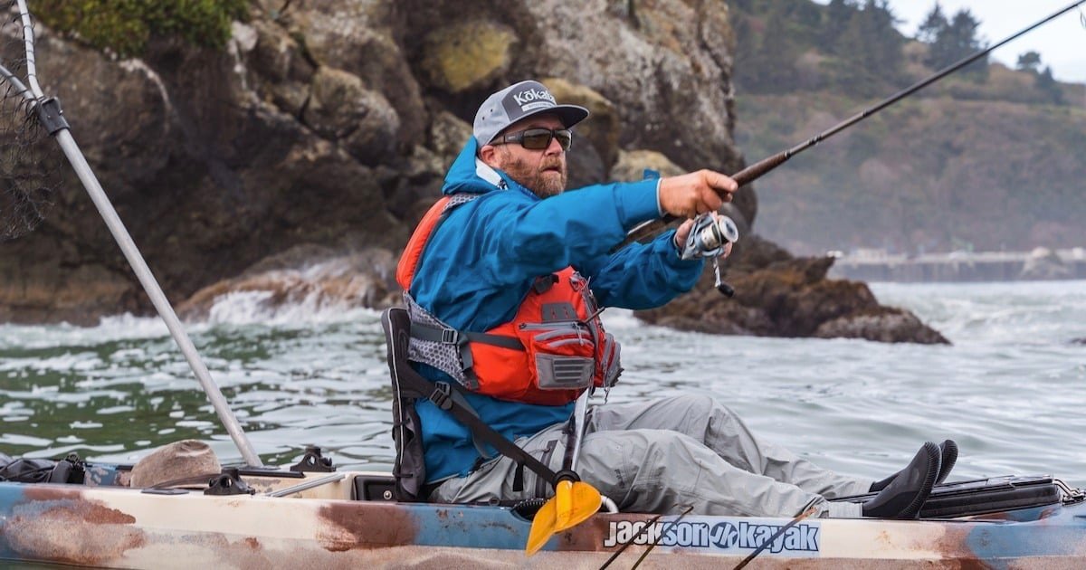 Person in a kayak wearing a blue jacket and orange life vest, fishing with a rod in a coastal water setting.