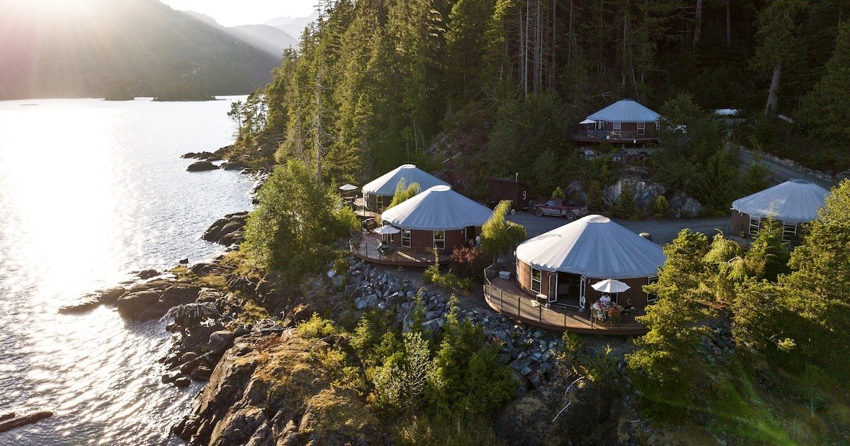 Three yurts situated near a body of water, surrounded by trees and rocky terrain, with sunlight reflecting off the water.