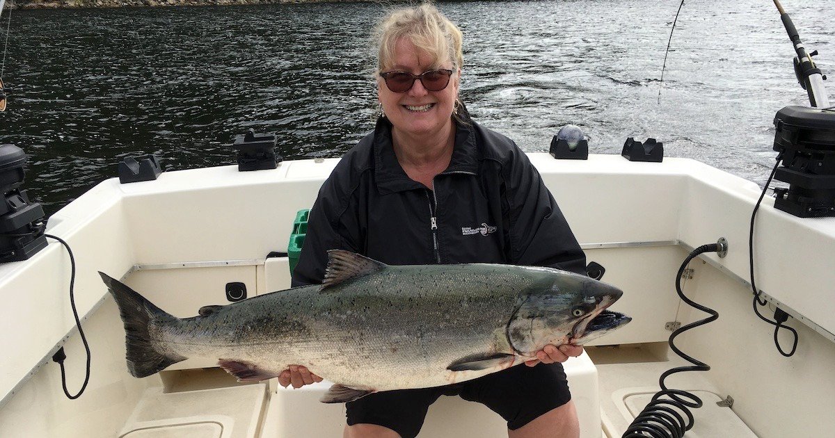Woman holding a large salmon while seated in a boat, wearing sunglasses and a black jacket.