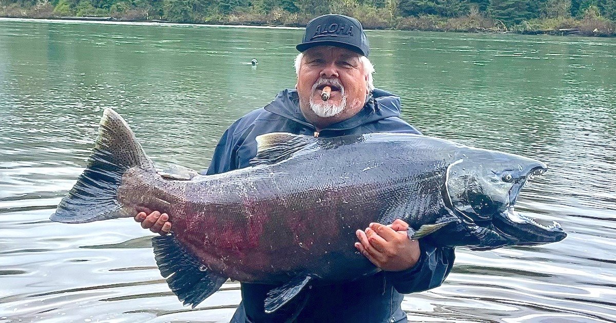 Man holding a large salmon fish while standing in water, wearing a black cap and smoking a cigar.