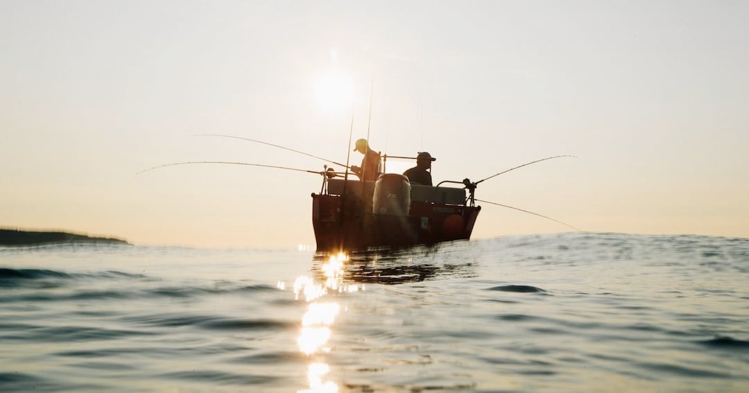 Fishing boat with two anglers casting rods into the water during sunrise, reflecting light on the surface.