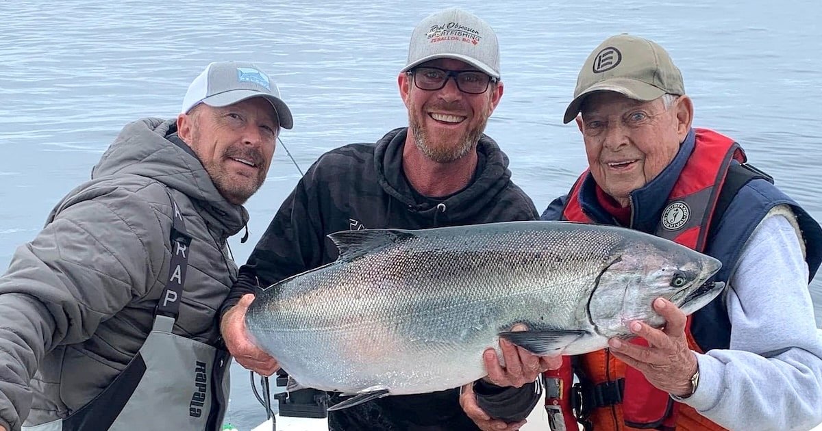 Three men holding a large salmon while posing on a fishing boat. The men are smiling, showcasing their catch.
