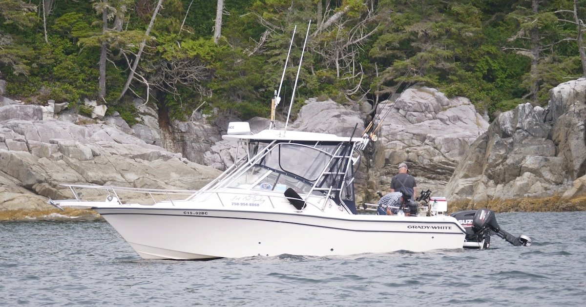 A fishing boat equipped with fishing rods and gear, navigating through calm waters near rocky shoreline vegetation.
