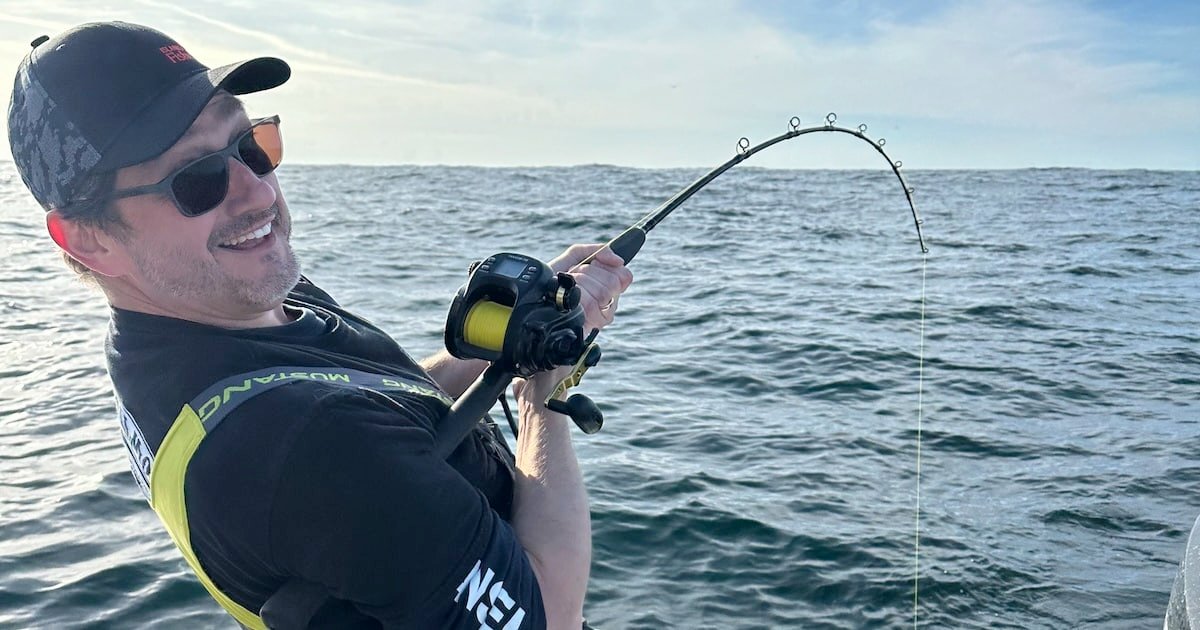 Person holding a fishing rod with a bent line, engaged in fishing on the water, wearing sunglasses and a cap.