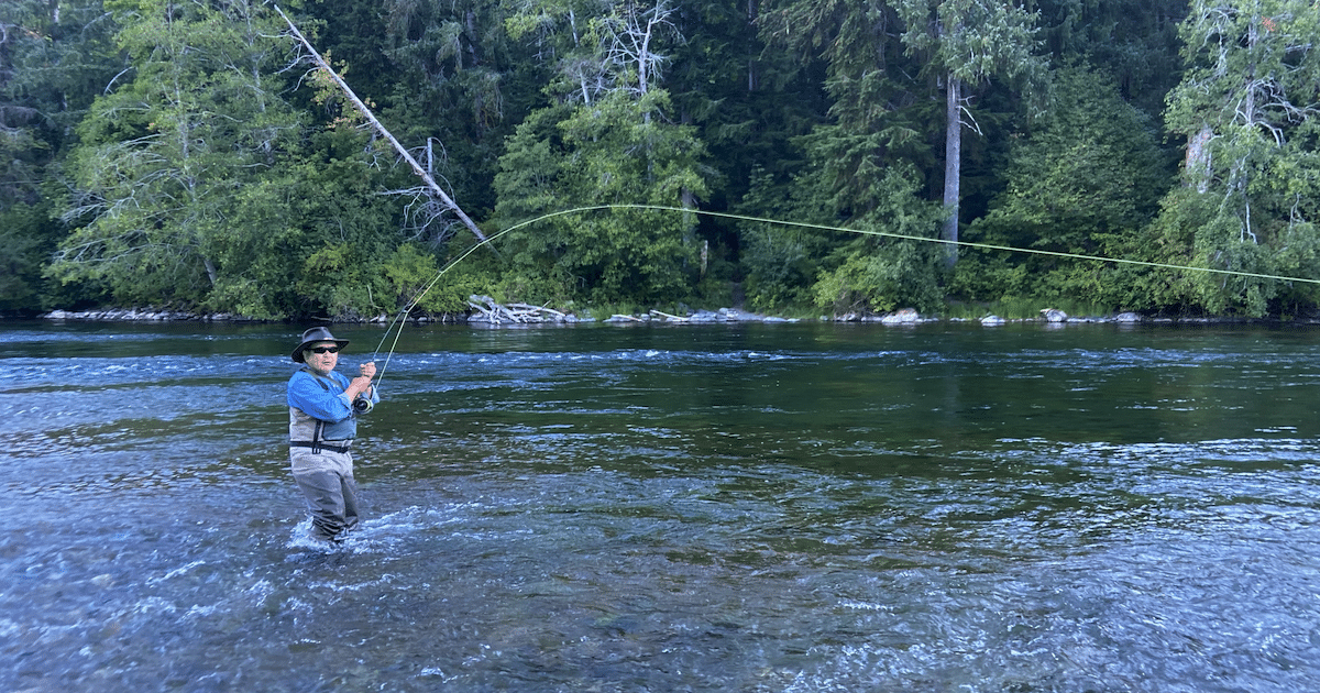 Angler standing in a river, casting a fishing line with a bent rod, surrounded by trees and water.