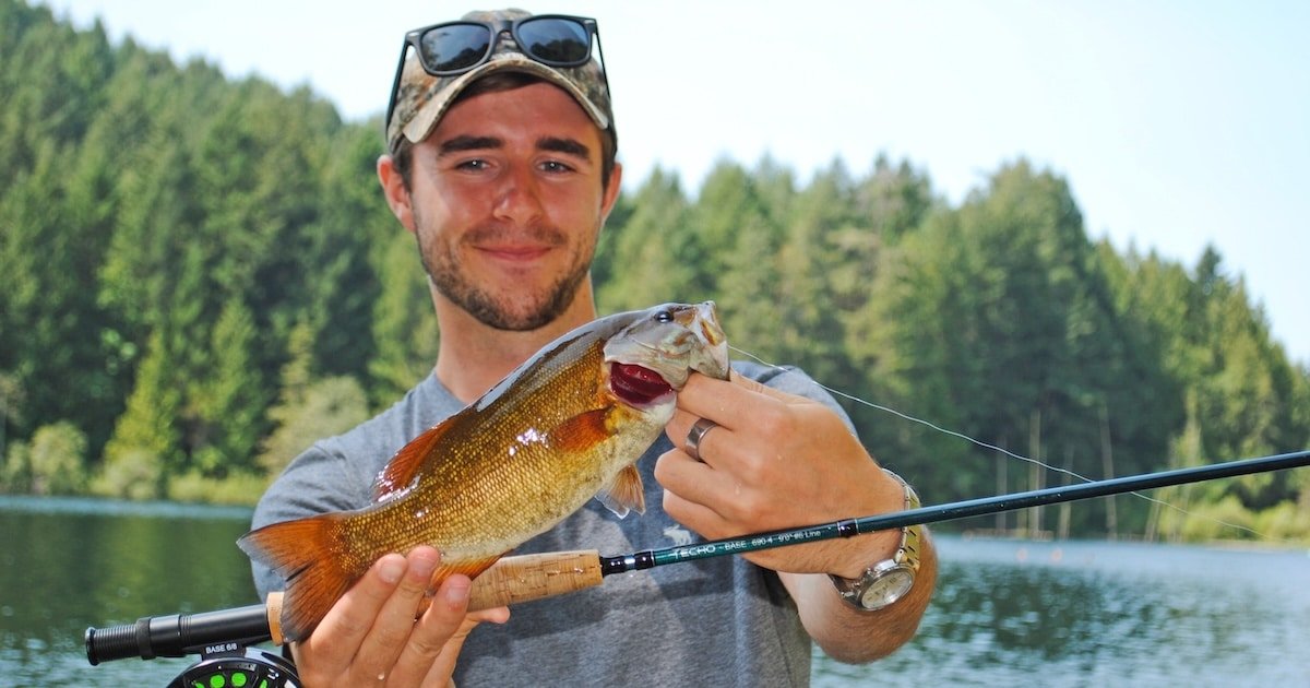 Man holding a bass fish while standing near water, wearing sunglasses and a camouflage hat, with a fishing rod in the foreground.