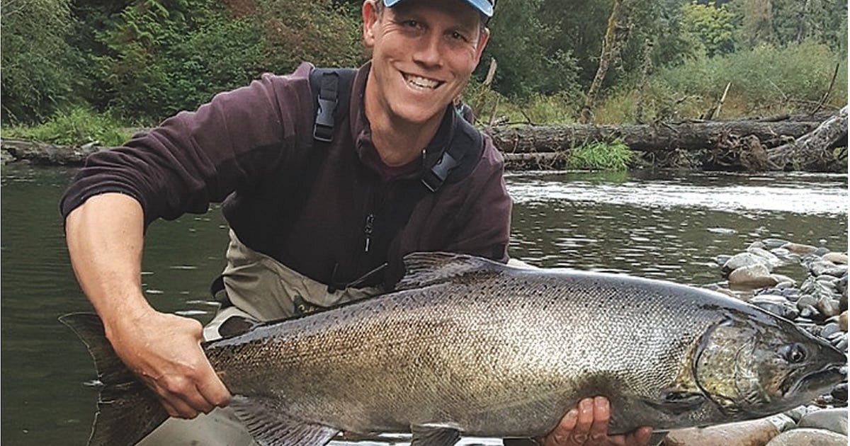 Person holding a large salmon fish while standing in a river, wearing fishing gear and a cap.