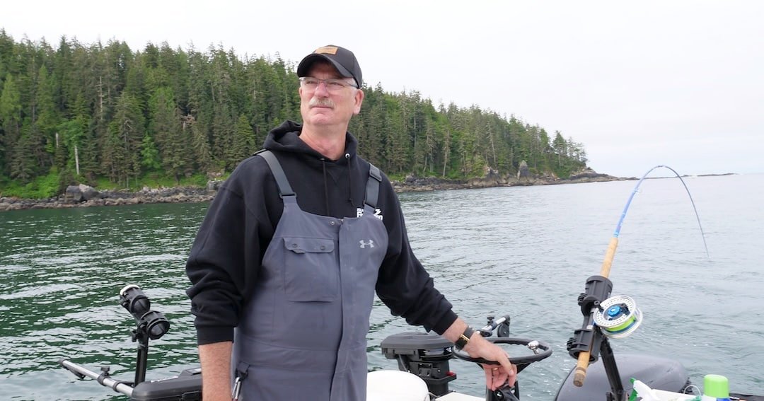 Man wearing a hoodie and fishing gear, standing on a boat with fishing equipment, surrounded by water and trees in the background.