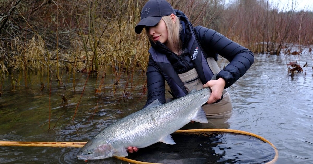 Woman holding a large fish while standing in shallow water, wearing fishing gear and a cap.