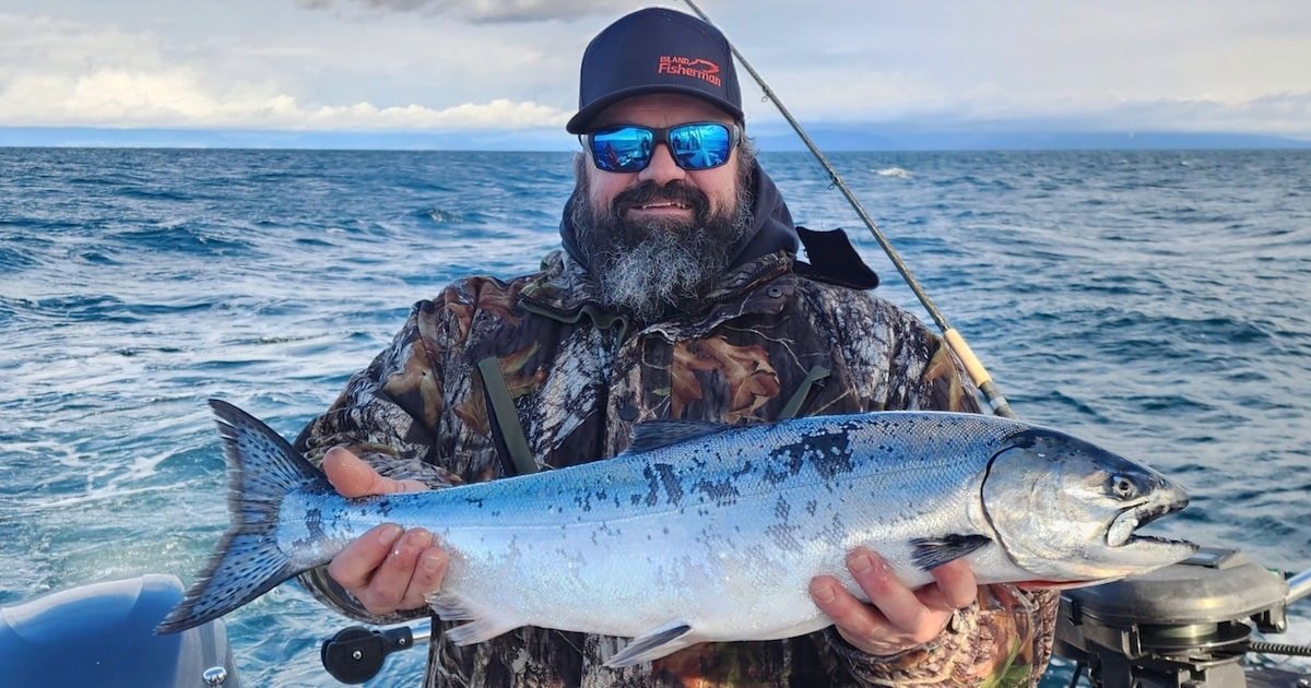Man holding a large salmon while wearing sunglasses and a camouflage jacket on a boat in the ocean.