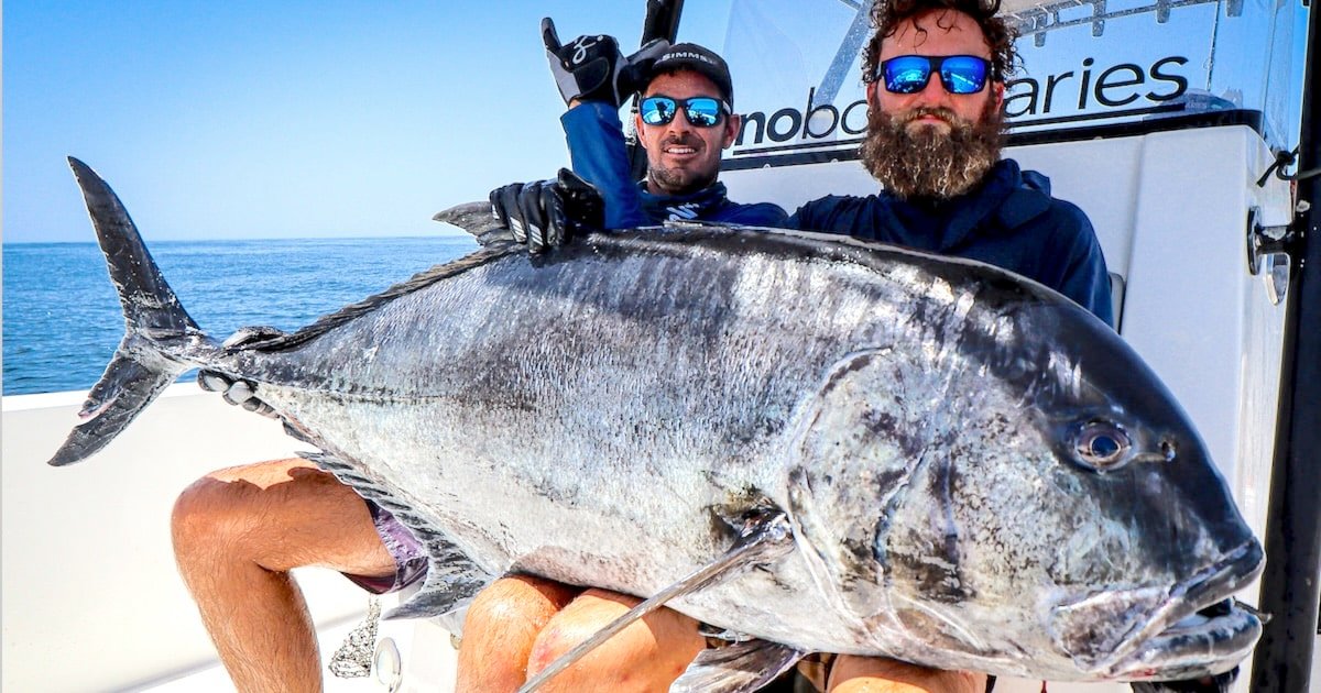 Two men holding a large fish on a boat, with one man making a hand gesture while the other poses with a beard and sunglasses.