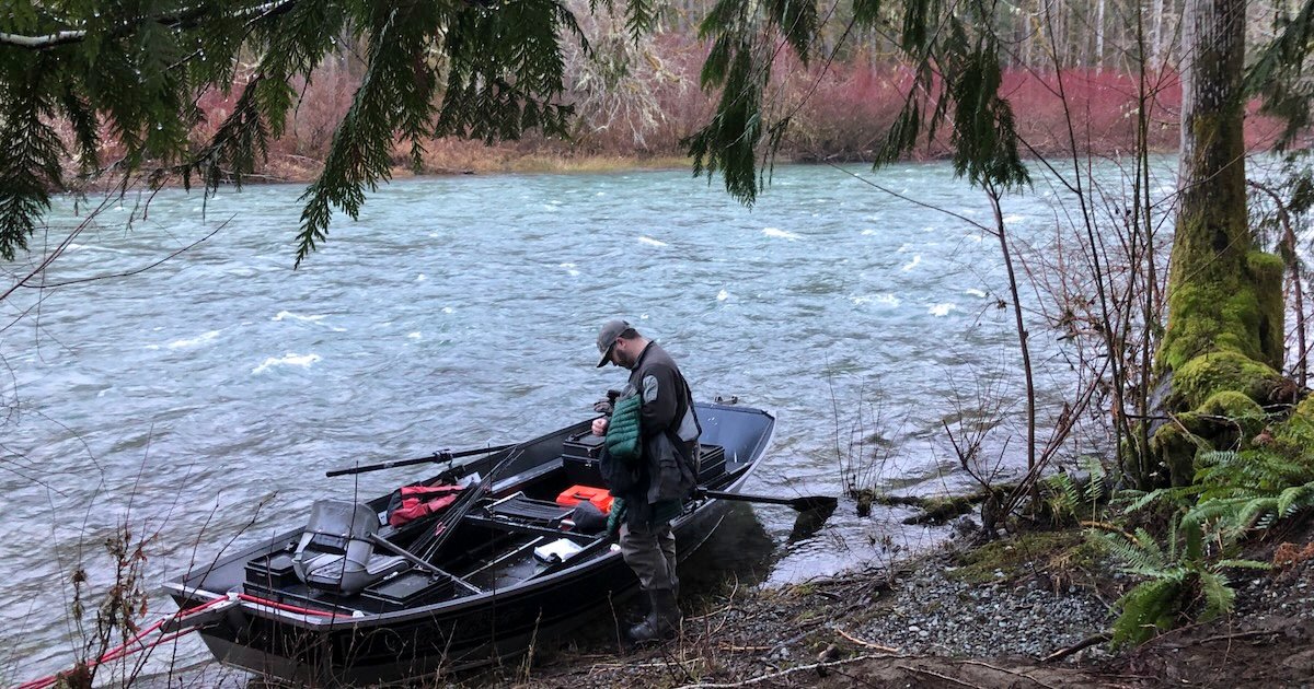 A person in a gray uniform standing beside a black boat on the bank of a river, preparing fishing equipment.