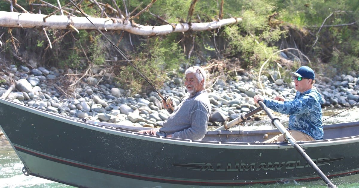 Two individuals in a fishing boat, one holding a fishing rod while the other rows, surrounded by natural scenery.