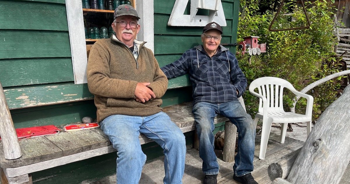 Two men sitting on a wooden bench outside a green building, both wearing casual clothing and hats.