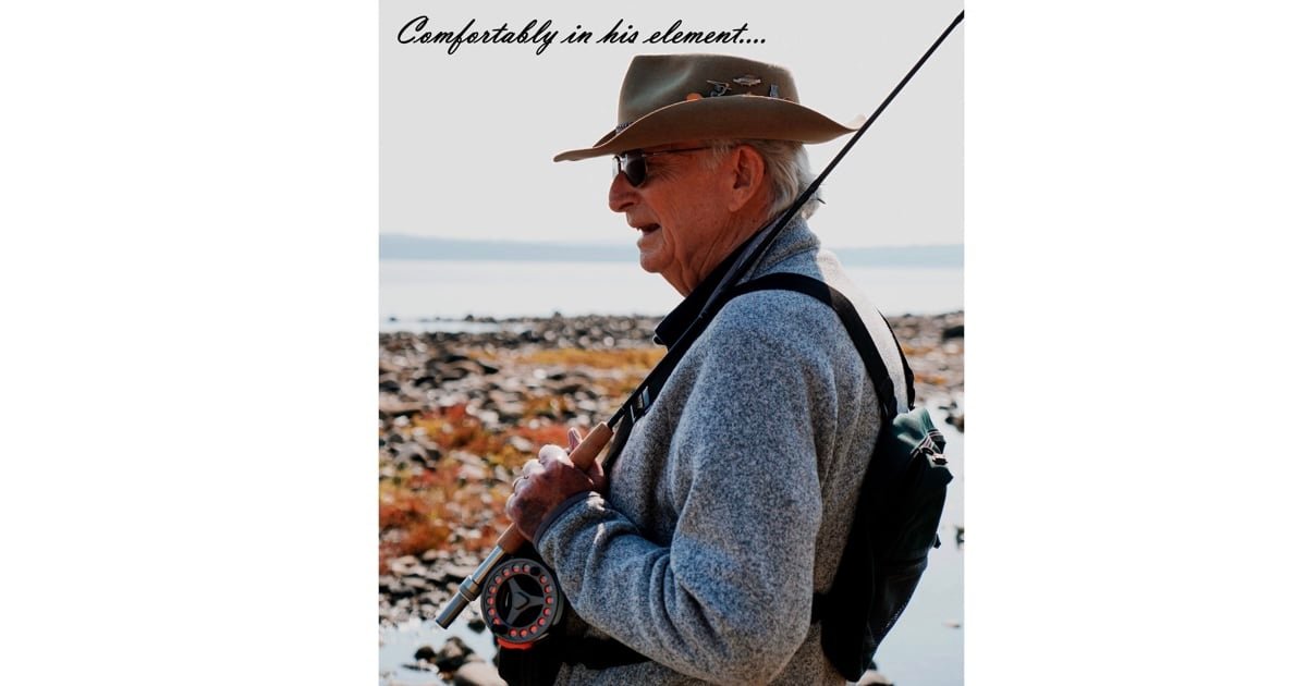 Elderly man wearing a hat and a sweater, holding a fishing rod, standing by the water's edge.