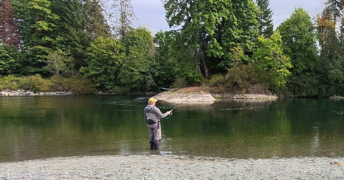 Person fishing in a river with trees and a rocky area in the background, casting a line into the water.