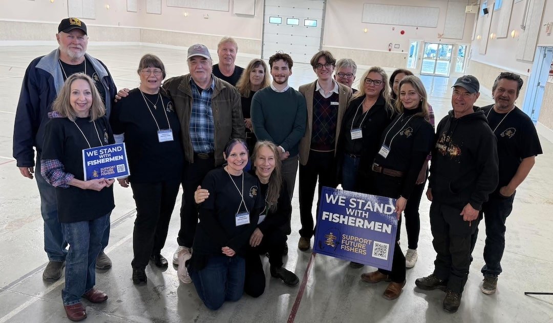 A group of fifteen BCRFA organizers and volunteers stand together in a large, empty hall holding "We Stand With Fishermen" signs following the public meeting.
