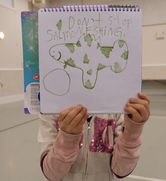 A child holds up a notebook drawing of a green fish with the handwritten text "Don't stop salmon fishing" at the Duncan town hall.