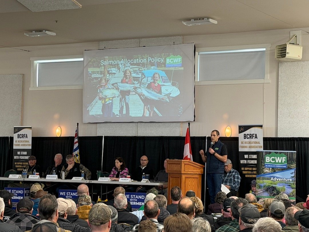 esse Zeman, Executive Director of the BC Wildlife Federation, addresses a capacity crowd at the Duncan town hall. A projector screen behind him displays "Salmon Allocation Policy" above a photo of children holding Chinook salmon.