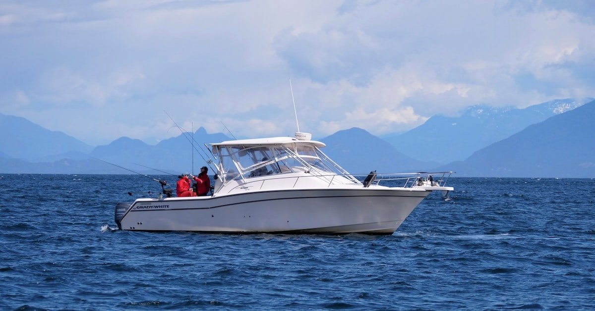 A white Grady-White walkaround fishing boat navigates the deep blue waters near Nanaimo, British Columbia. Two anglers in red waterproof gear are positioned at the stern, tending to multiple trolling lines. The backdrop features the dramatic, layered blue silhouettes of the Coast Mountains under a bright, cloud-dappled sky. Will the Salmon Allocation Policy Change this way of life?